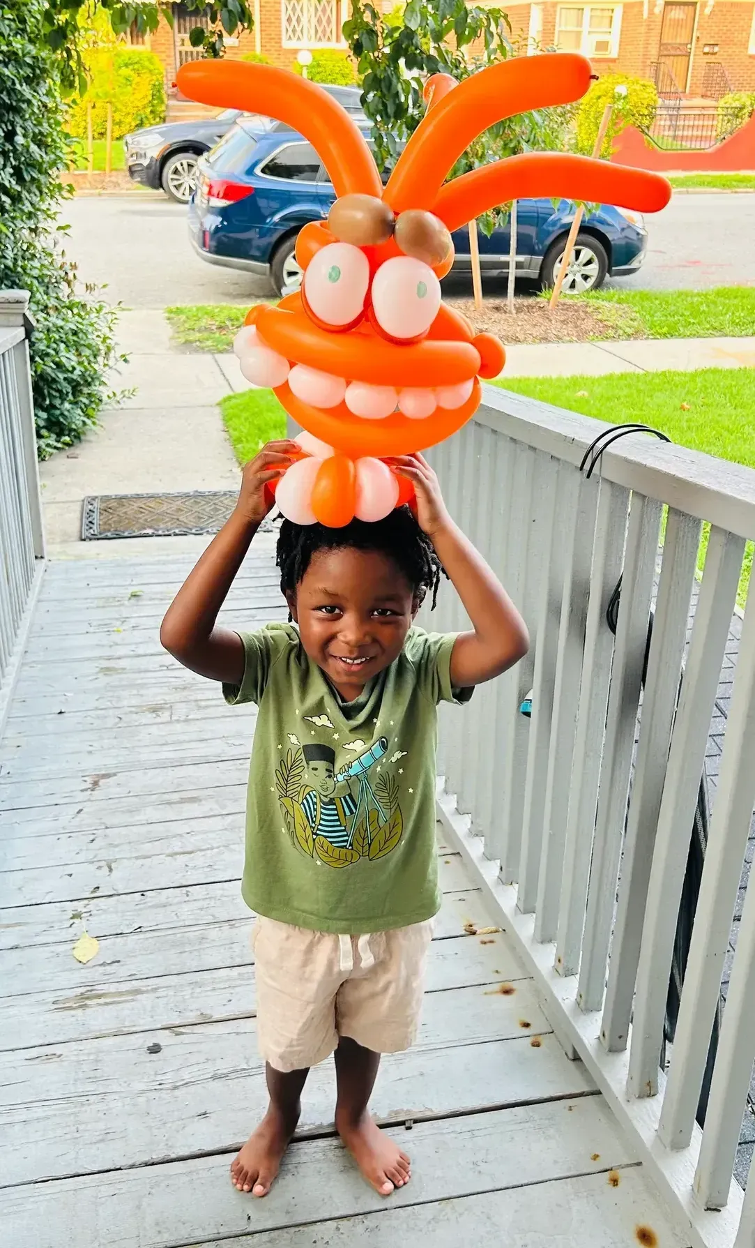 Child holding a colorful orange balloon creature on their head, smiling on a porch.