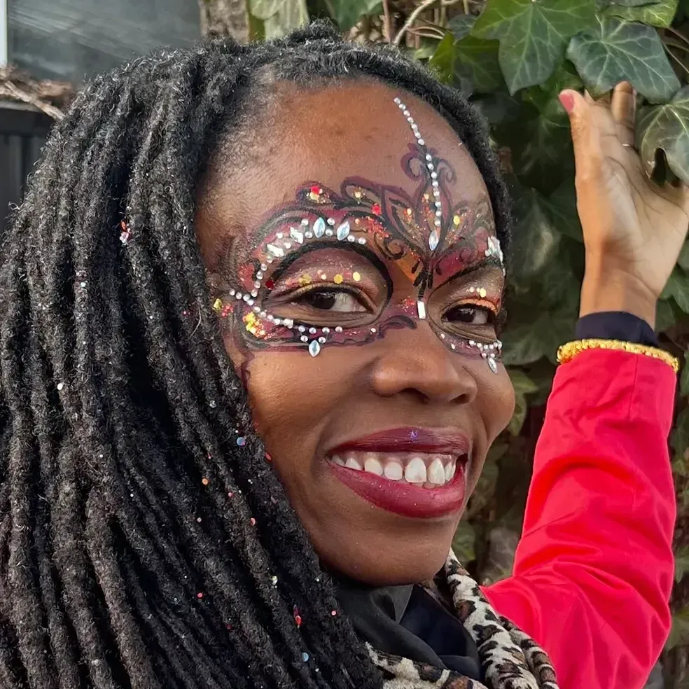 Woman with face paint, smiling, reaching toward ivy. She wears a red jacket and has dreadlocks.