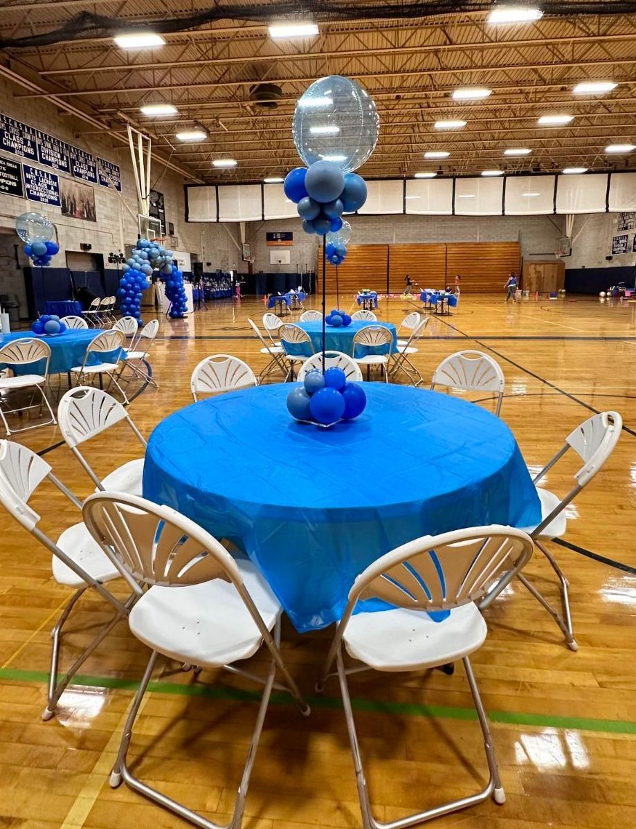 Tables with blue tablecloths and white chairs in a gymnasium, decorated with blue balloons.