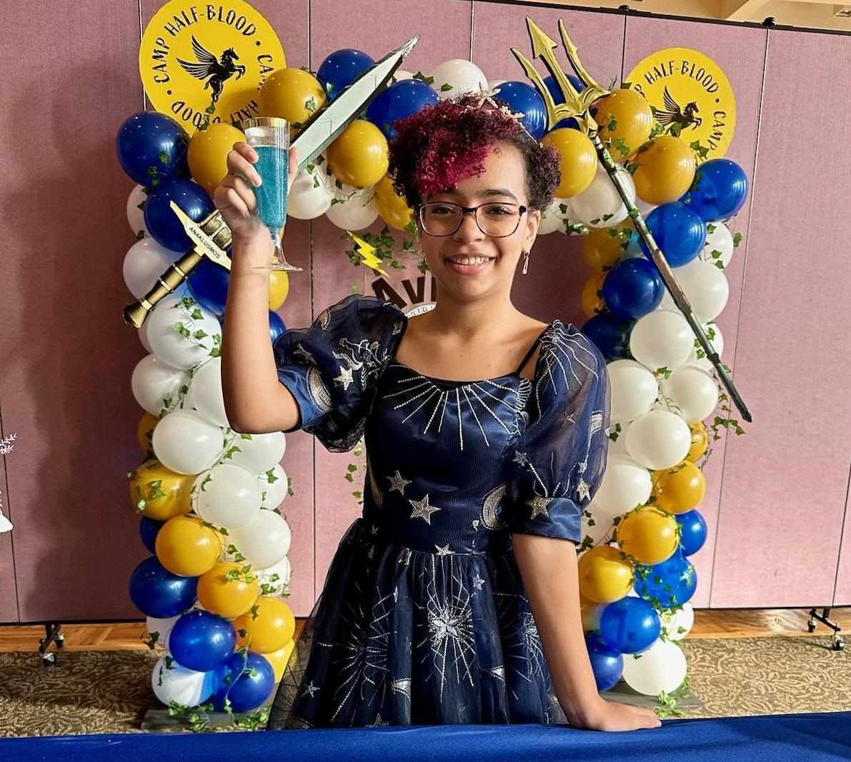 Person smiling, holding blue drink, standing before arch of balloons. Wearing starry dress, surrounded by decorations.