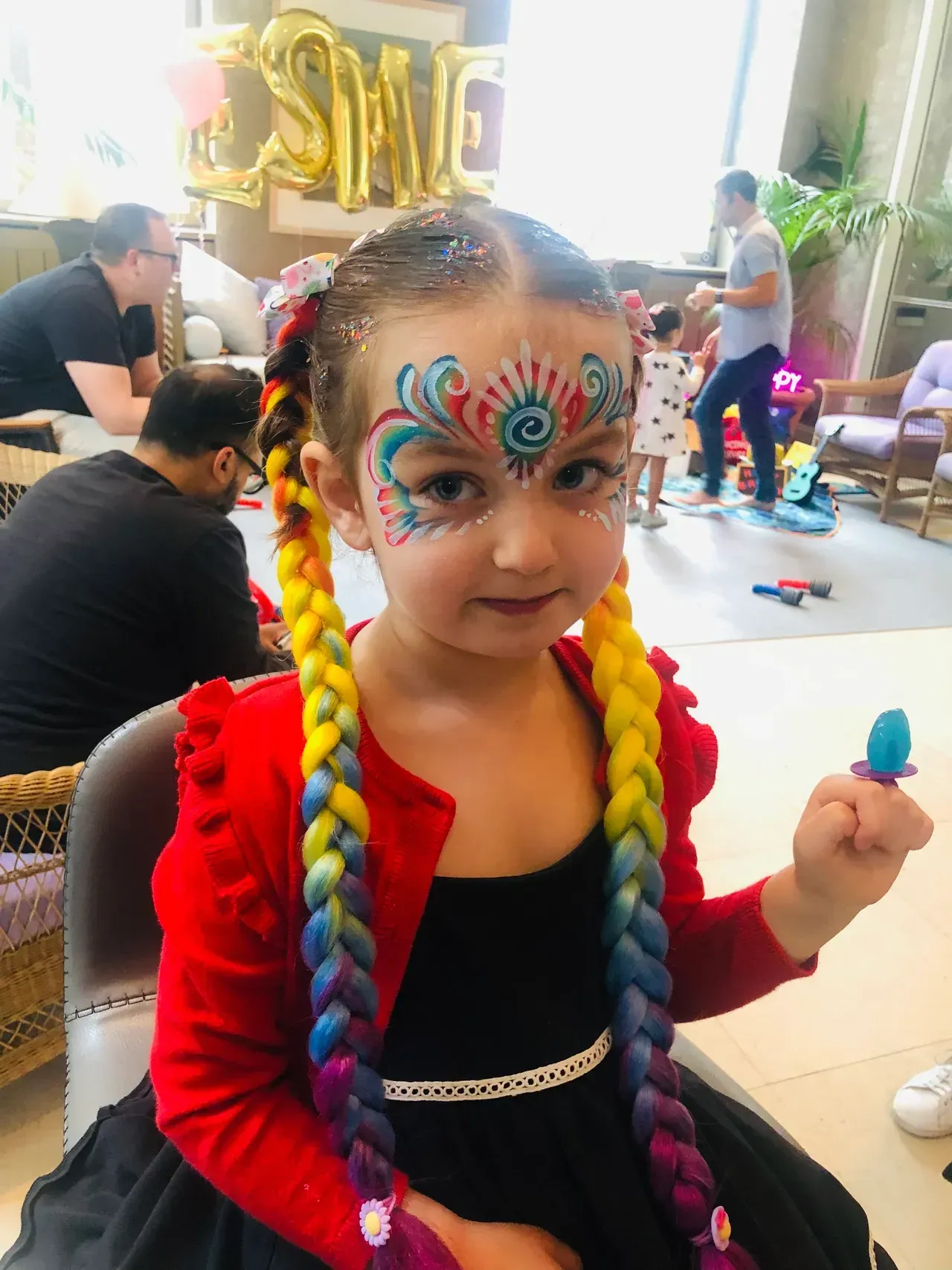 Girl with rainbow braids and face paint at a party, holding a toy ring pop.