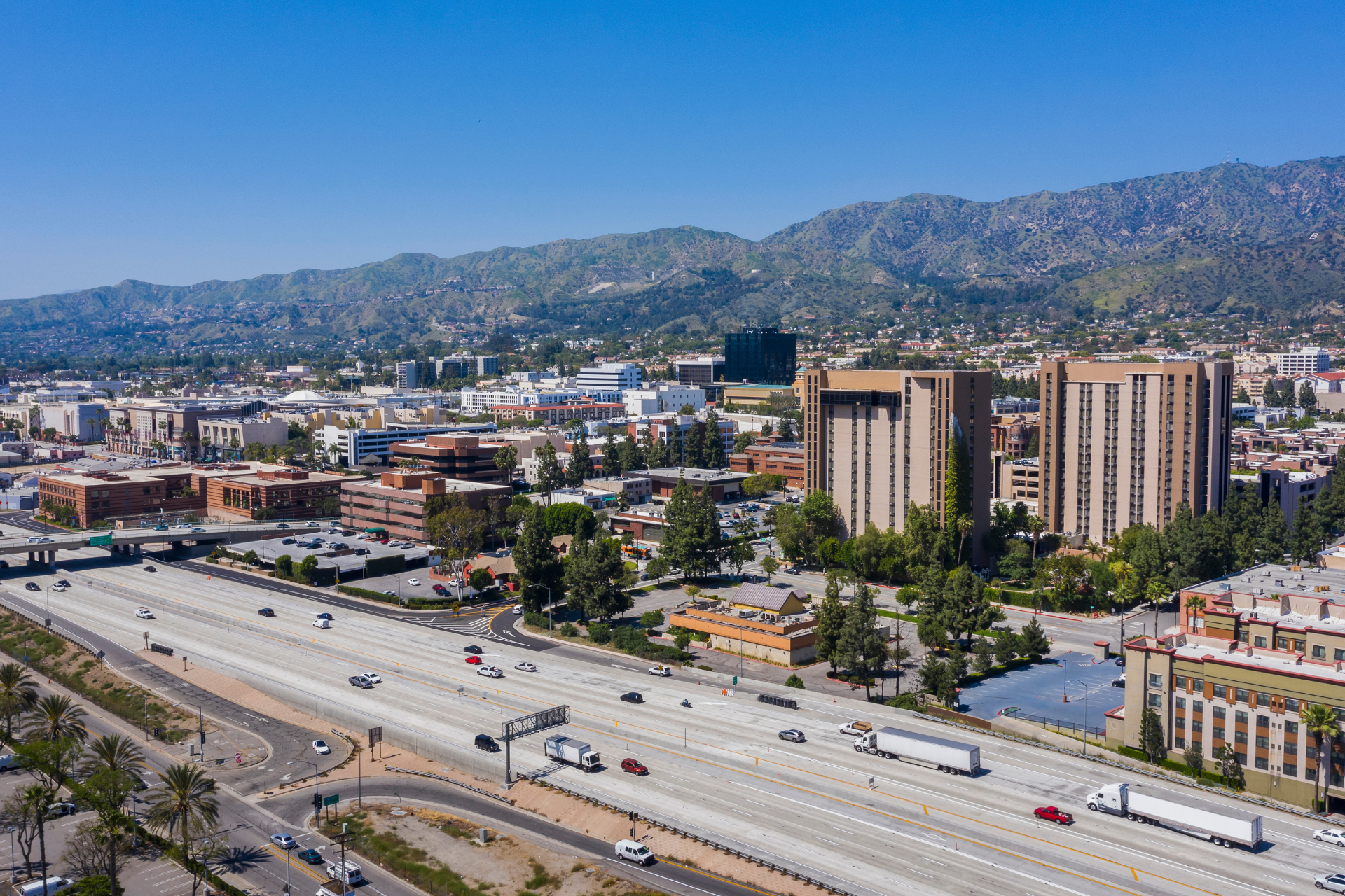 Cityscape with highway in foreground, buildings, and mountains under a blue sky.