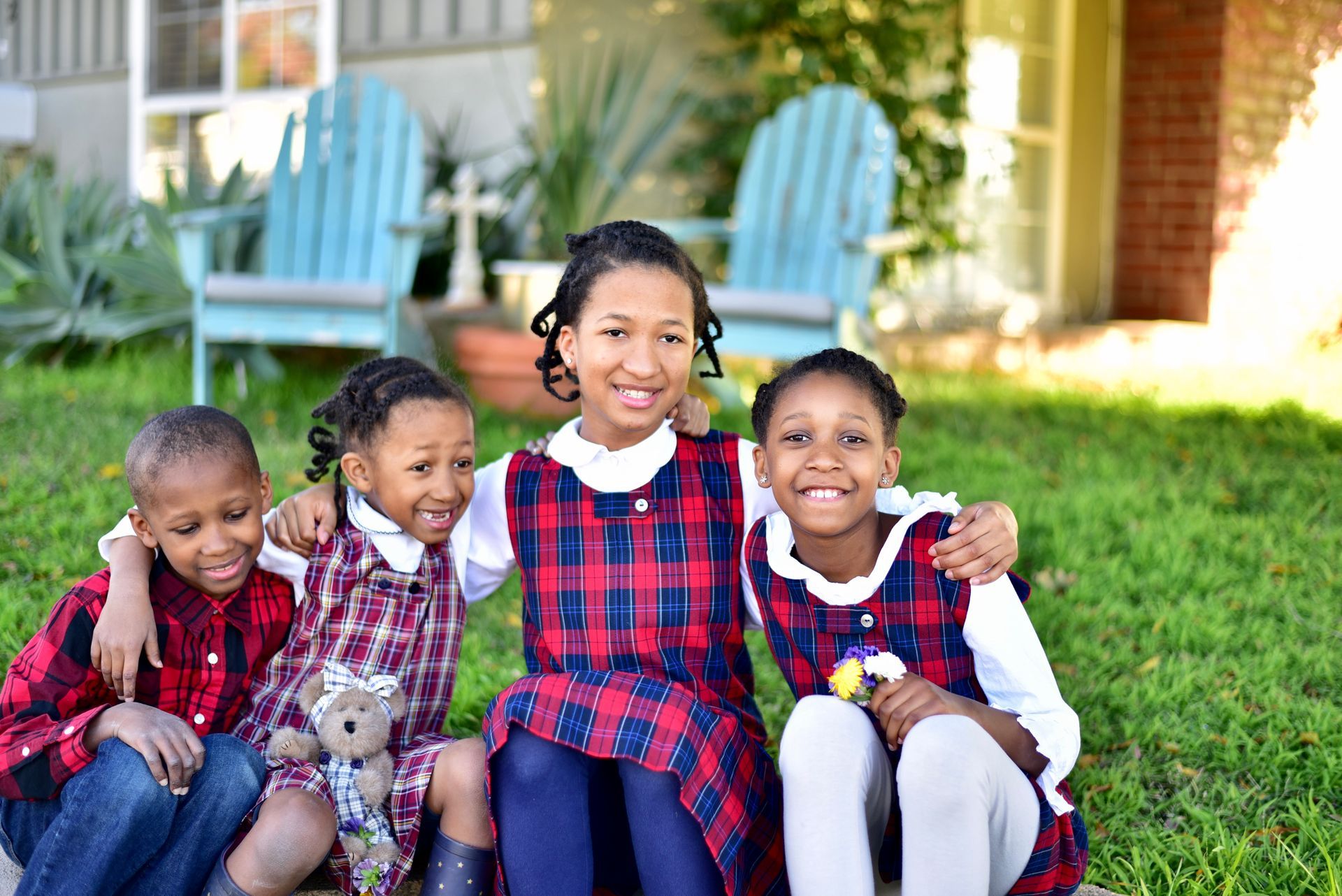Four children sitting together on grass, smiling in front of a house with blue chairs.