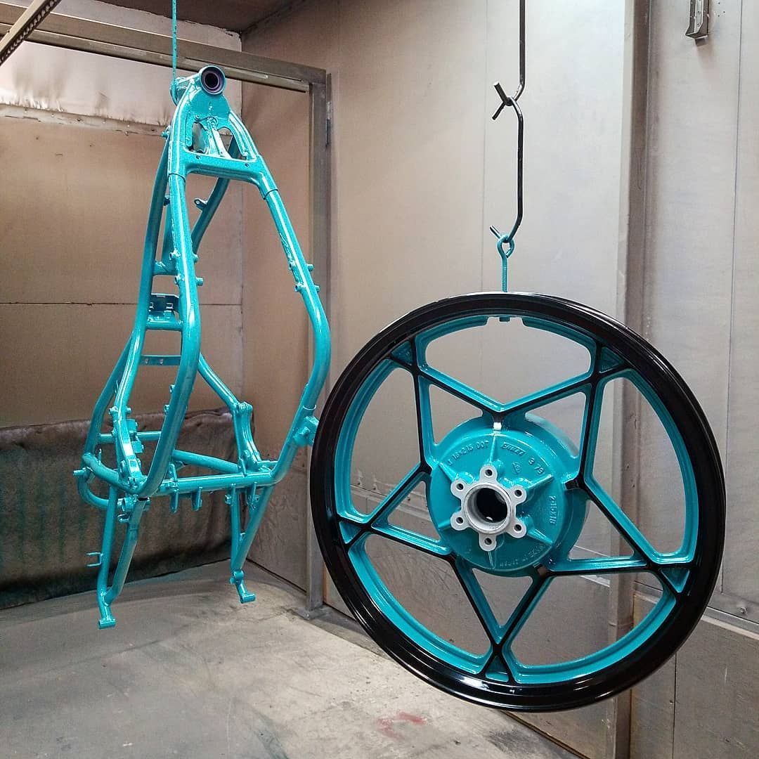 A motorcycle frame and wheel, both aqua blue with black accents, hang in a spray booth.