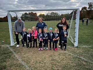 Group of children and adults posing in front of a soccer goal on a grassy field.