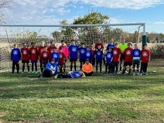 Youth soccer team posing in front of a goal, with players wearing red, blue, and orange jerseys.