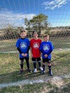 Three children in red and blue soccer jerseys standing on a field in front of a goal net