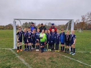 Youth soccer team posing on a field in front of a goal, with medals and coaches behind them.