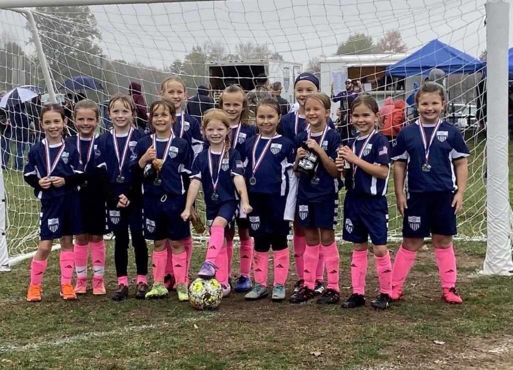 Youth soccer team posing in medals and navy uniforms on a rainy field in front of a goal.