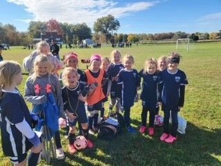 Youth soccer team posing on a grassy field with pink balls, wearing navy uniforms and orange pinnies.