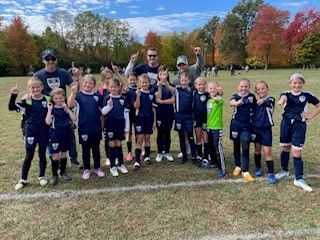 Youth soccer team posing on a grassy field, wearing navy uniforms with autumn trees in the background.