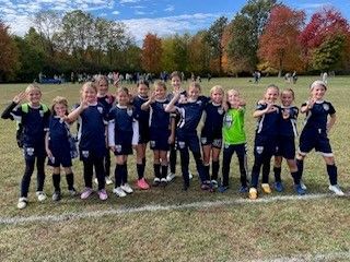 Youth soccer team posing on a grassy field, smiling in navy uniforms with trees in autumn behind them.