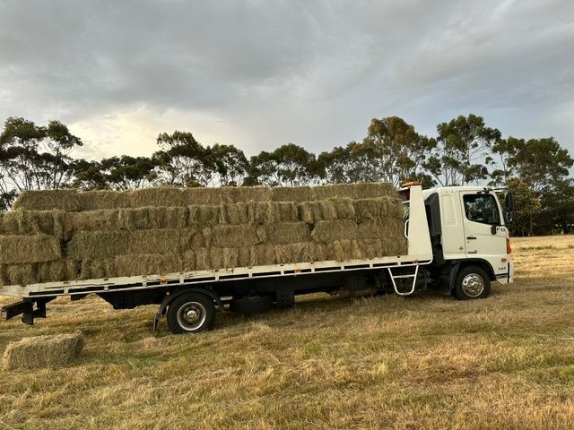 White flatbed truck loaded with hay bales in a dry field under a cloudy sky— Westside Panels & Towing in Prospect Vale, TAS
