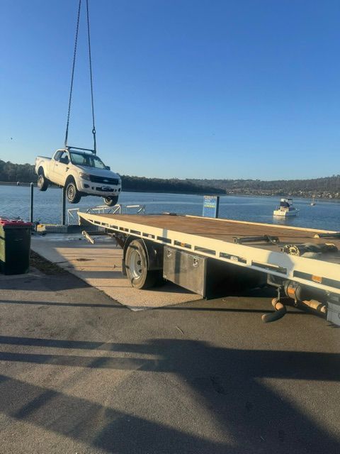 White SUV suspended by crane above a flatbed trailer near a waterfront dock at sunset — Westside Panels & Towing in Prospect Vale, TAS
