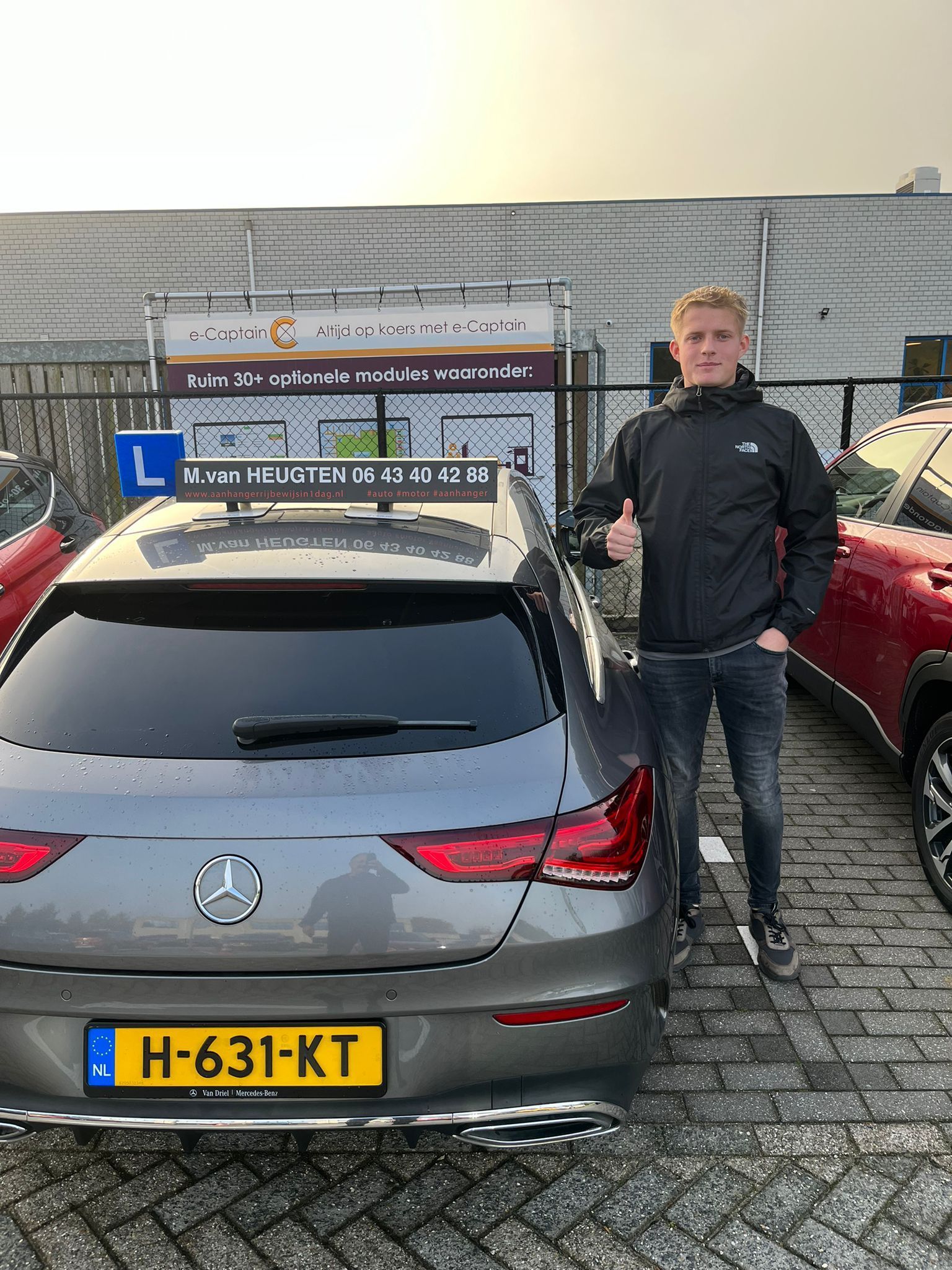 Een man staat naast een grijze Mercedes met een dakkoffer en een Nederlands kenteken op een parkeerplaats.