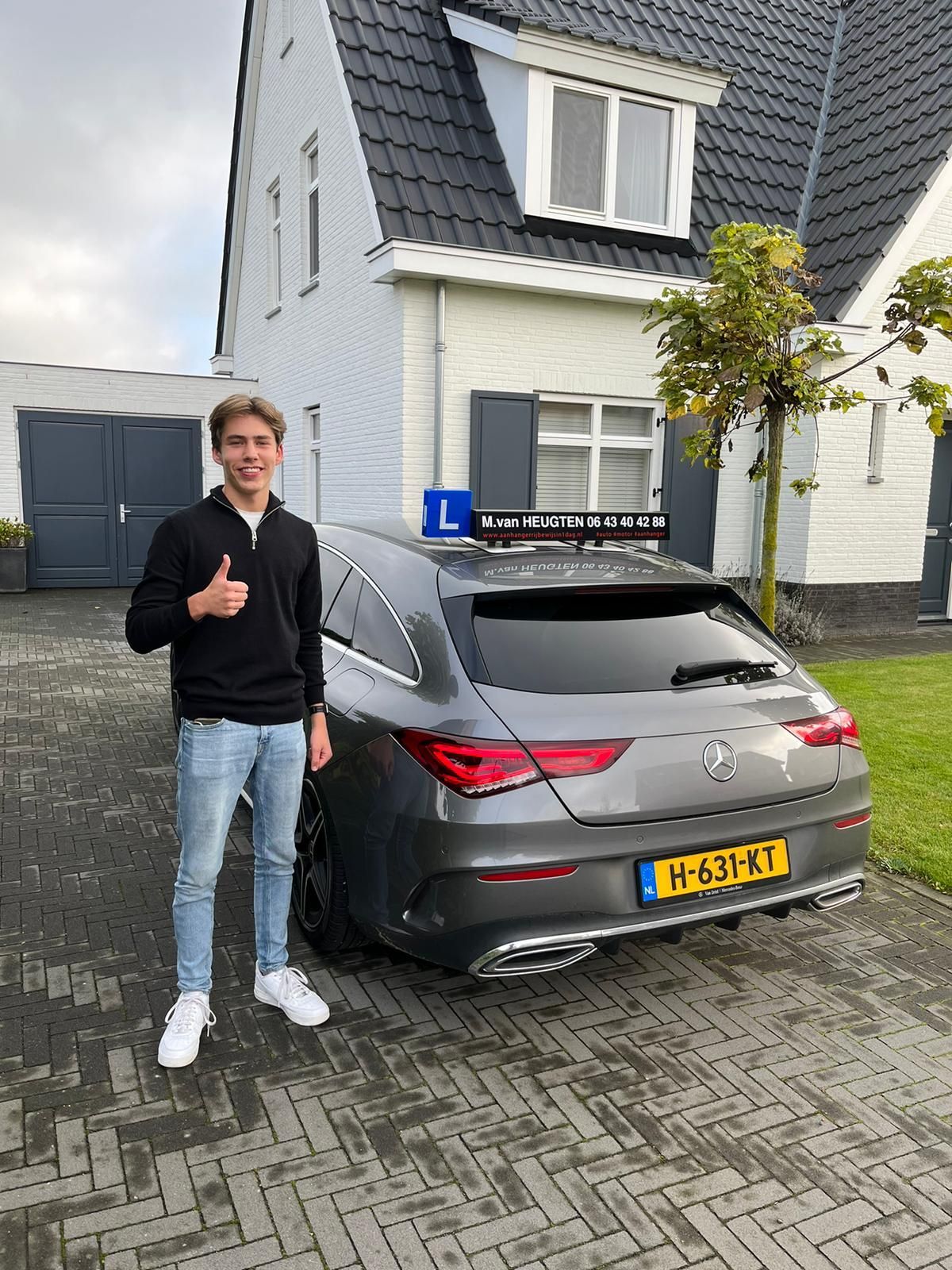 Een persoon staat naast een grijze auto voor een wit huis en steekt zijn duim omhoog.