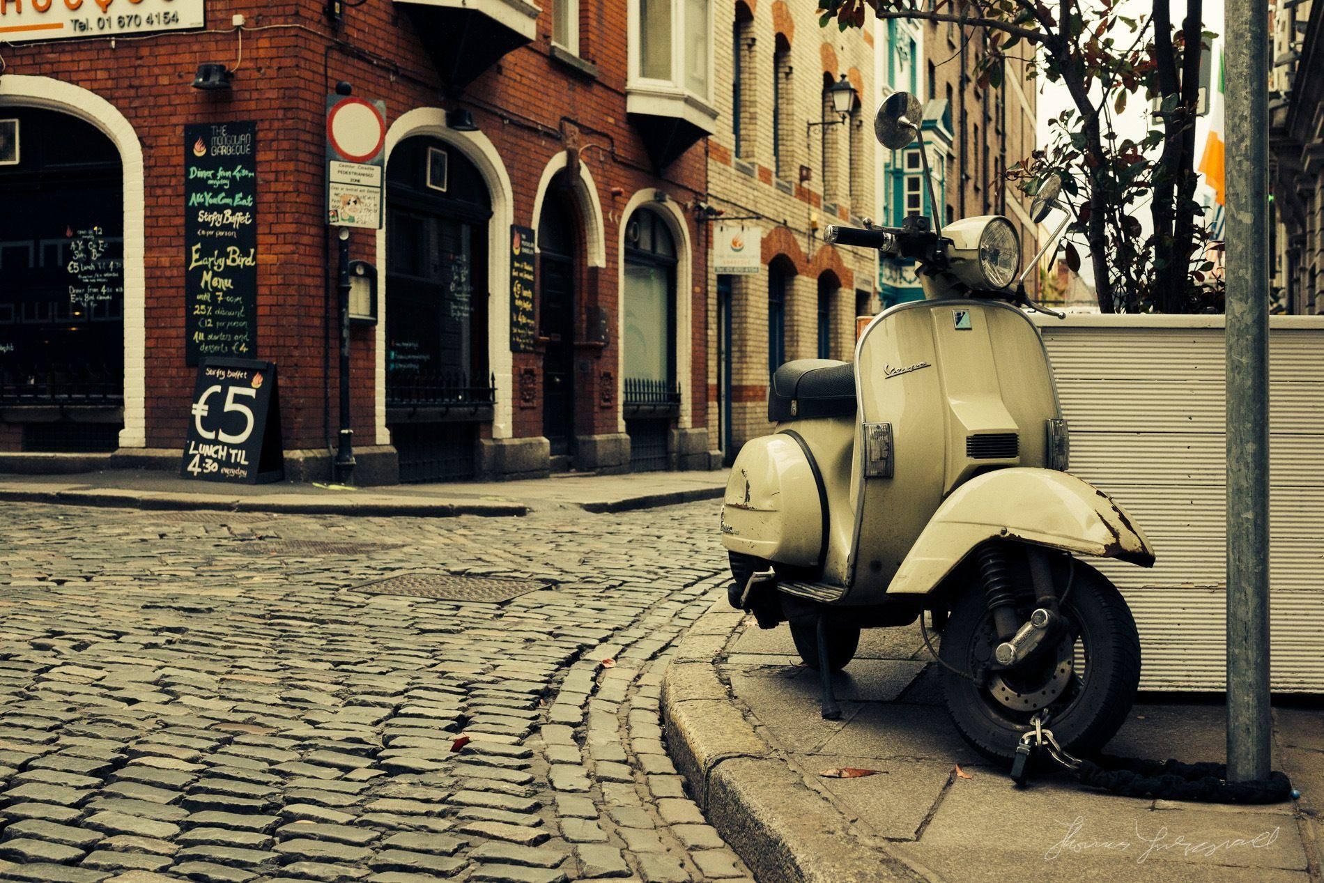 Beige vintage scooter geparkeerd op een straat met kinderkopjes voor oude bakstenen gebouwen.