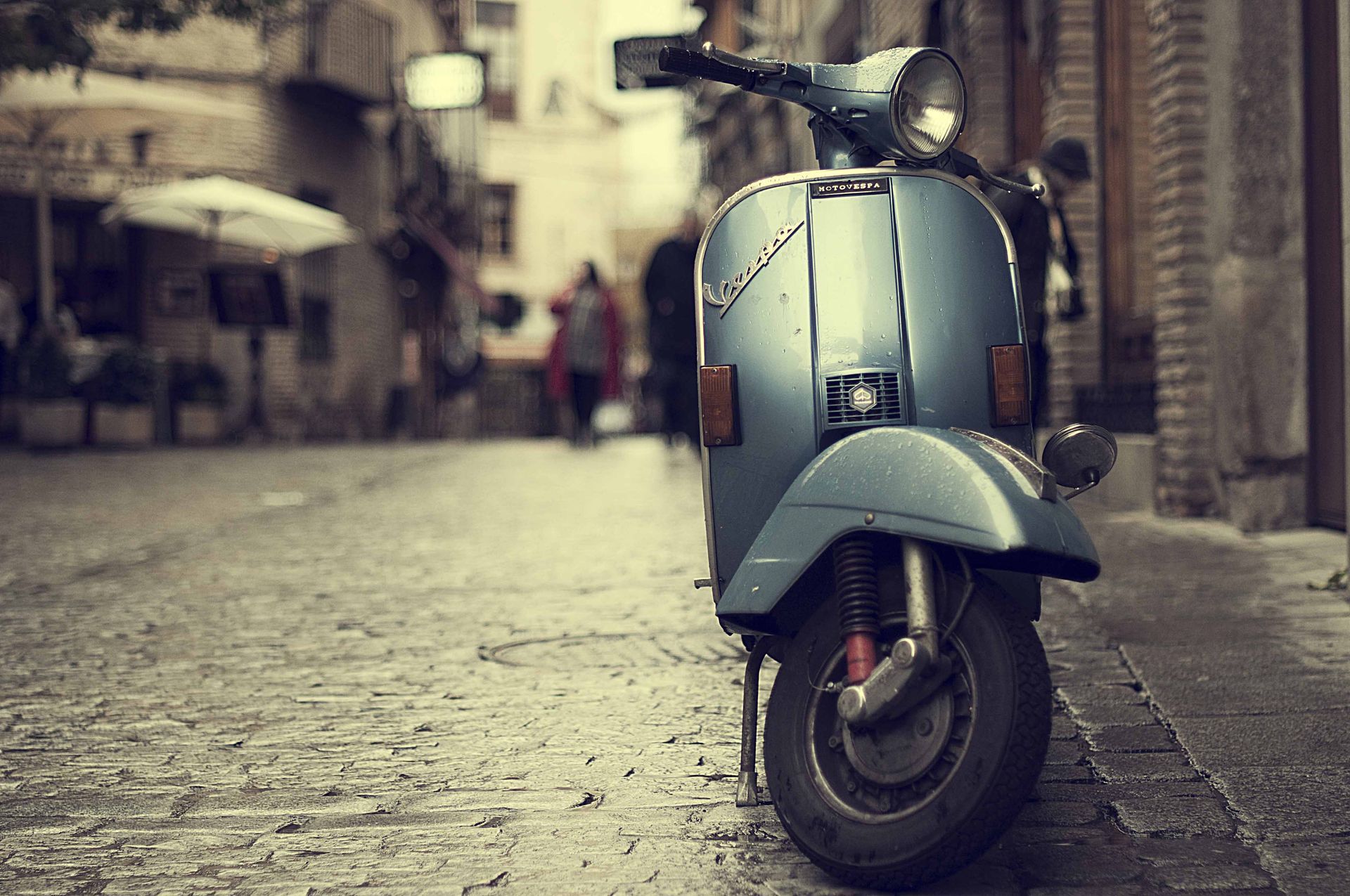 Een blauwe vintage scooter staat geparkeerd op een straat met kinderkopjes in een wazige, oude stadsomgeving.