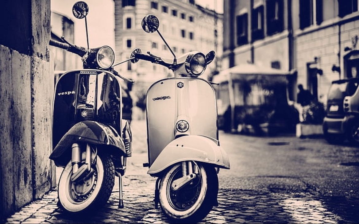 Een vintage scooter geparkeerd in een smalle stadsstraat, gefotografeerd in blauw zwart-wit.