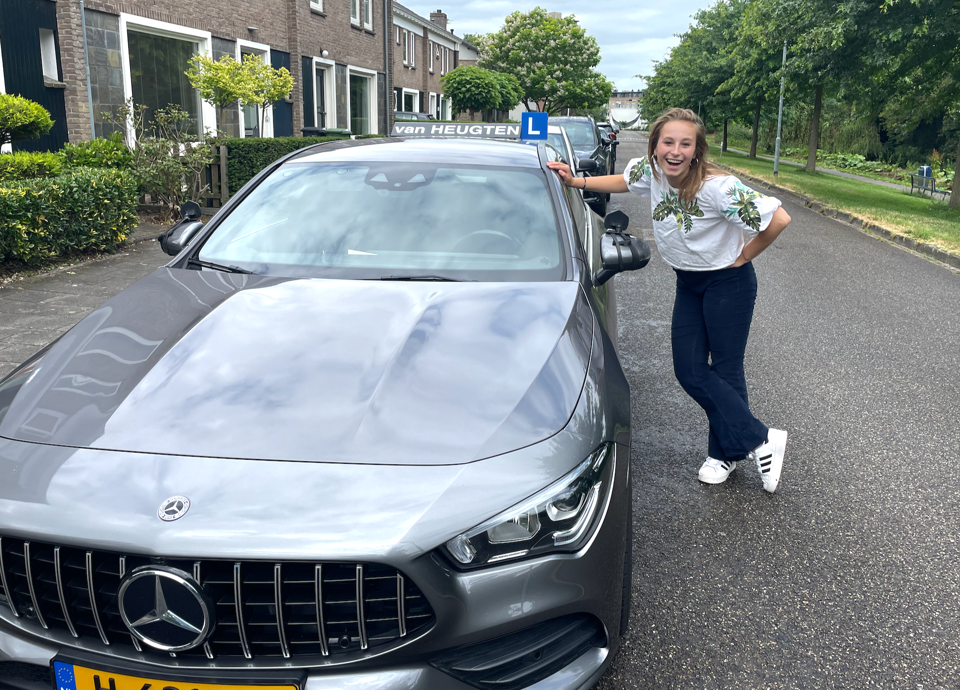 Vrouw poseert naast een grijze Mercedes in een woonwijk.