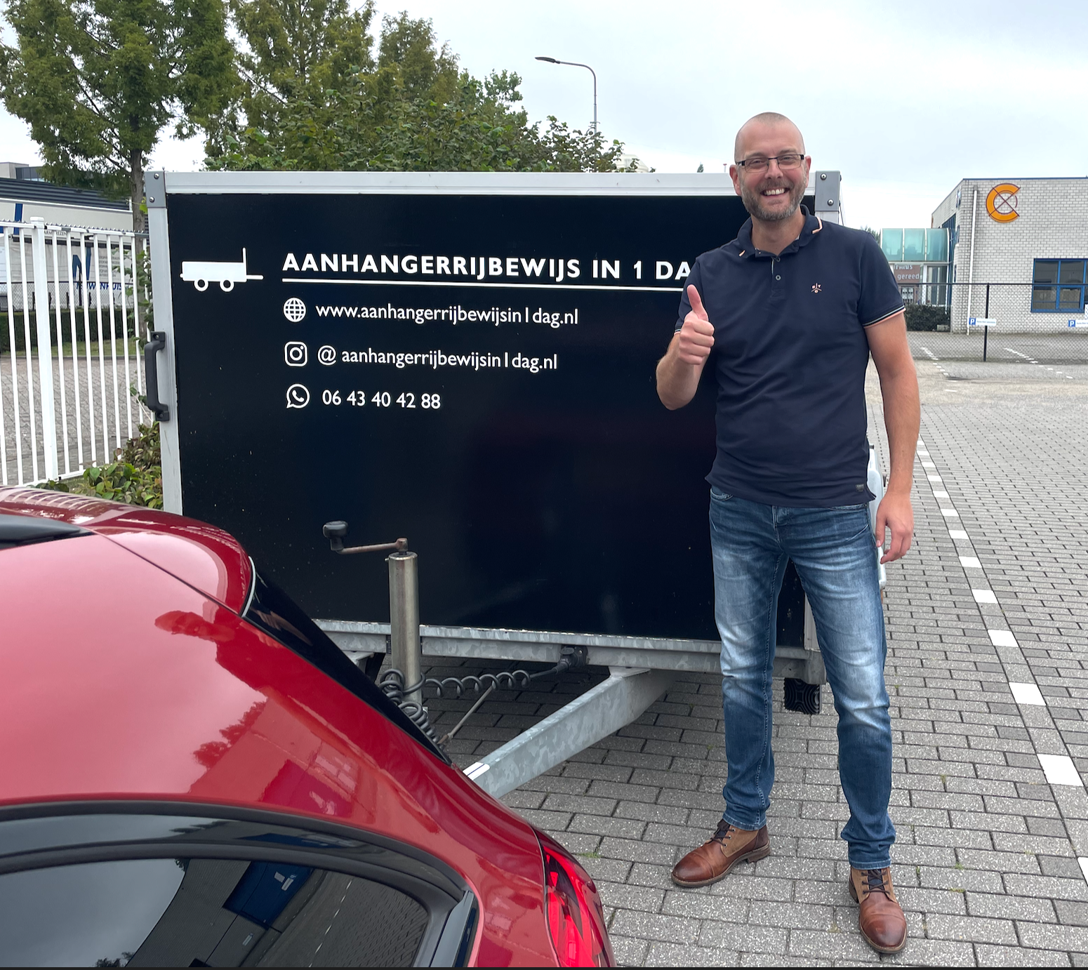 Een man staat naast een zwarte aanhanger met witte tekst, naast een rode auto op een verhard terrein.