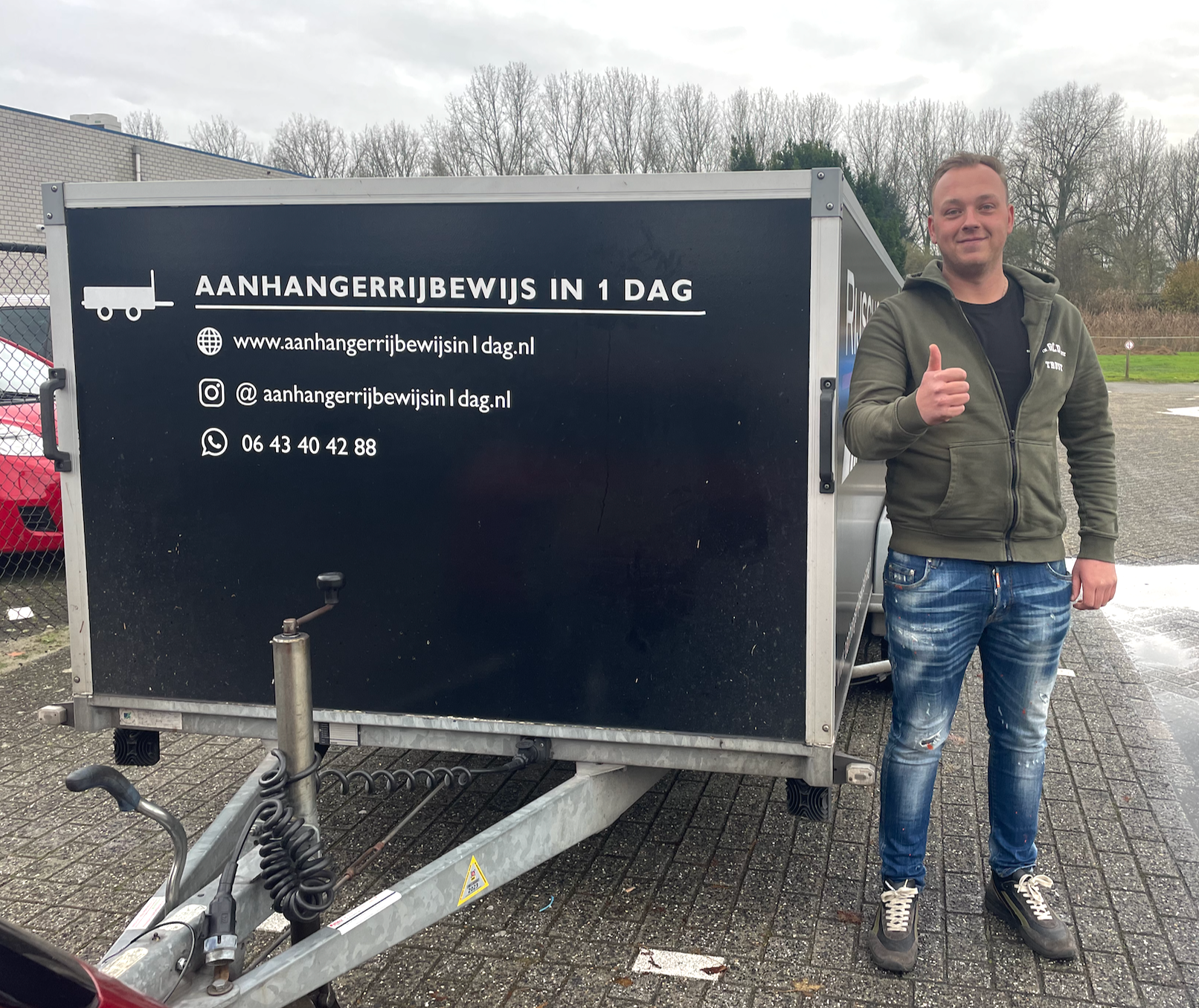 Een man staat naast een zwarte aanhanger met Nederlandse tekst en steekt zijn duim omhoog in de buitenlucht op een bewolkte dag.