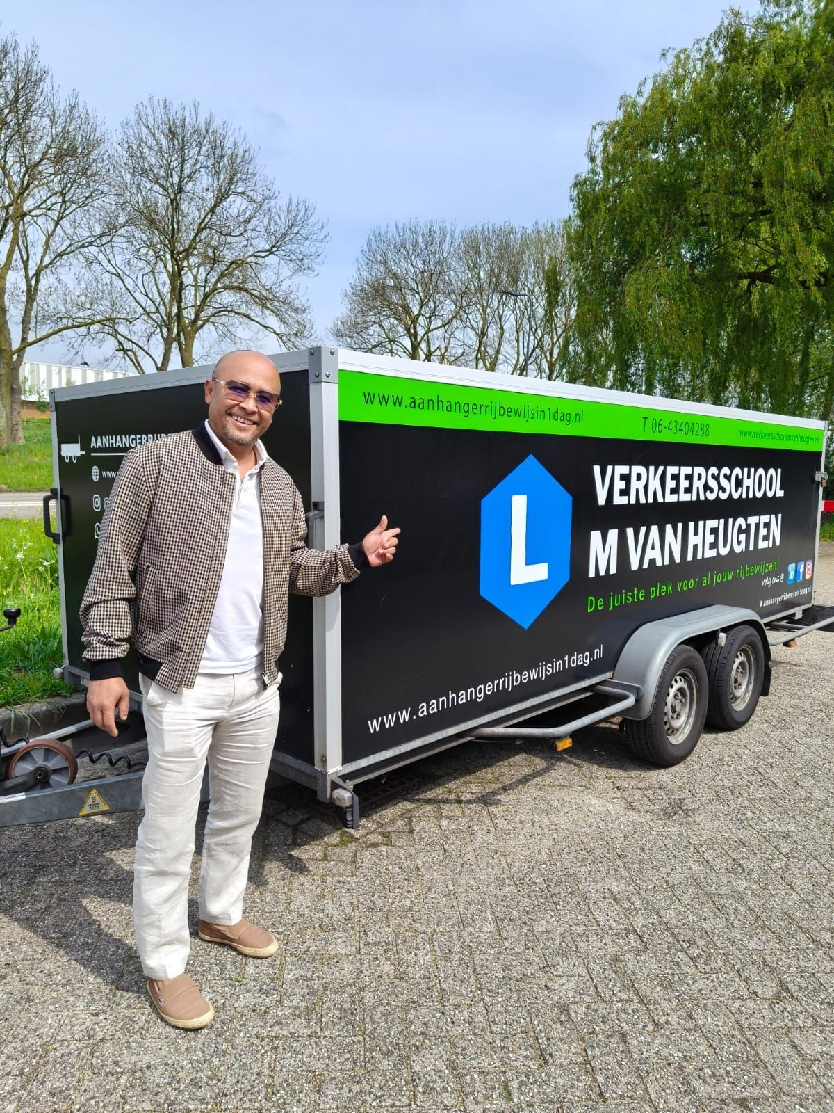 Een man staat naast een zwarte trailer met het logo van een Nederlandse school op een grindterrein buiten.