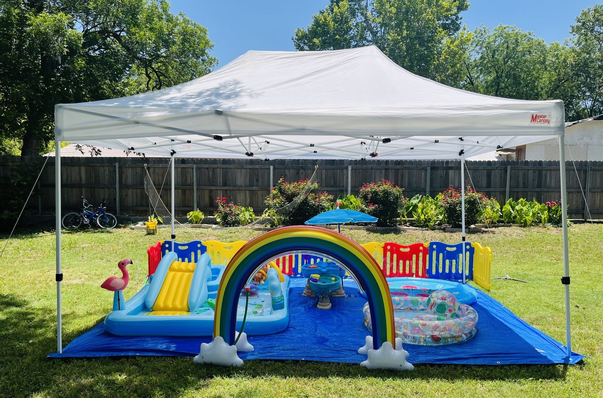 A large inflatable pool with a rainbow and a slide under a white tent in a backyard.