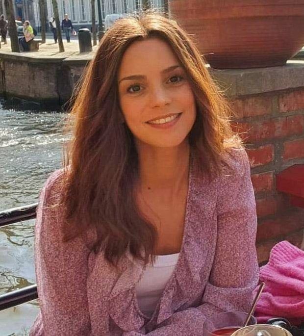Vrouw in een roze top lacht naast een bakstenen muur langs de rivier, met water en boten op de achtergrond.