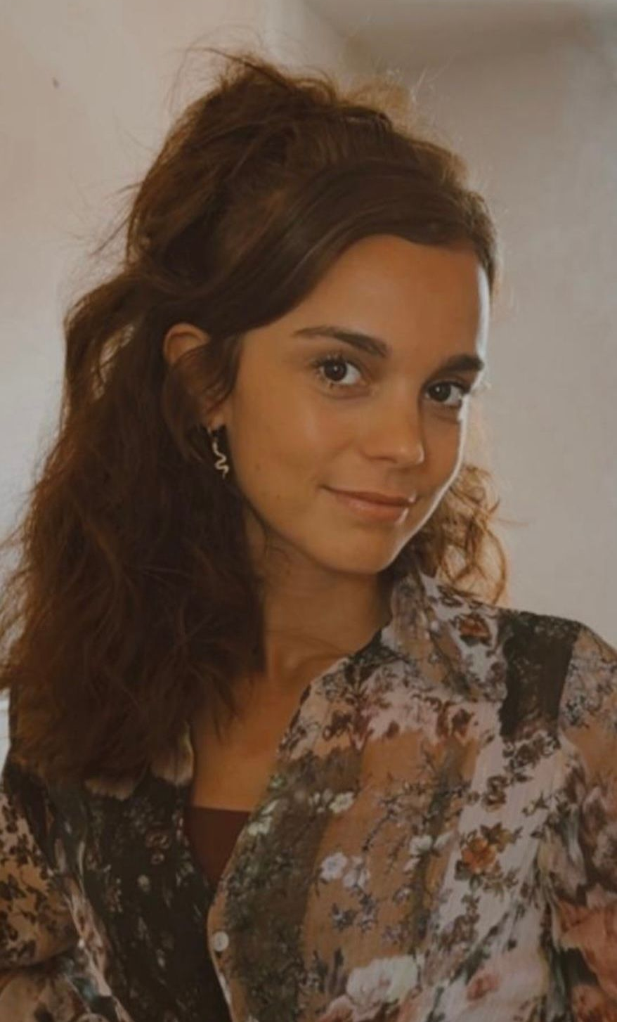 Portret van een lachende vrouw met krullend haar, gekleed in een bloemenblouse, tegen een lichte achtergrond.