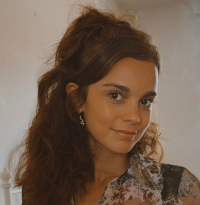 Vrouw met bruin haar glimlacht zachtjes in een bloemenblouse tegen een effen binnenachtergrond.