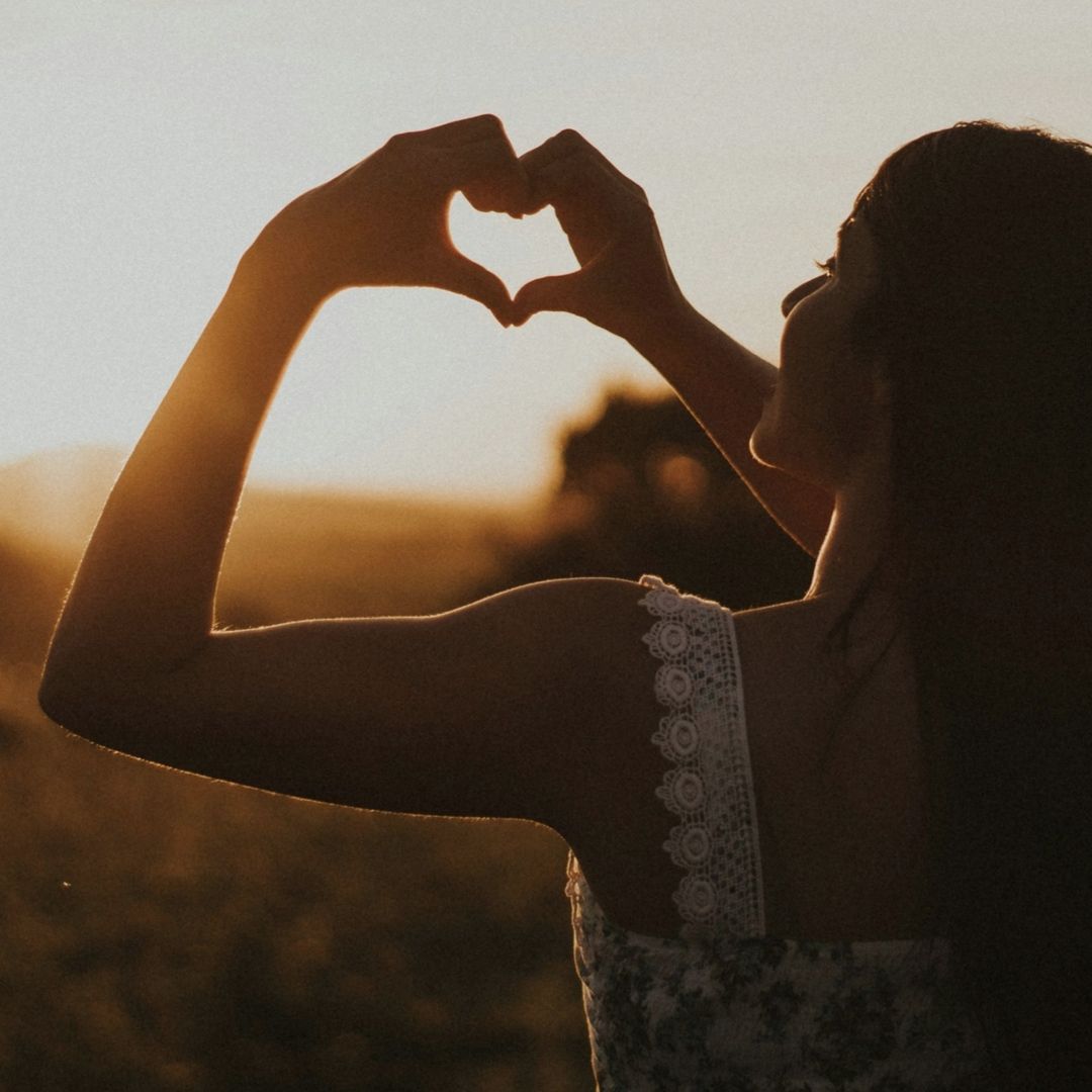 Een vrouw die met haar handen een hart vormt, afgetekend tegen een achtergrond van de ondergaande zon in een veld.