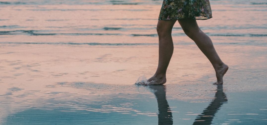 Een persoon loopt op blote voeten op een nat strand, waarbij de weerspiegeling van de benen, de kleding en de zonsondergang zichtbaar is.