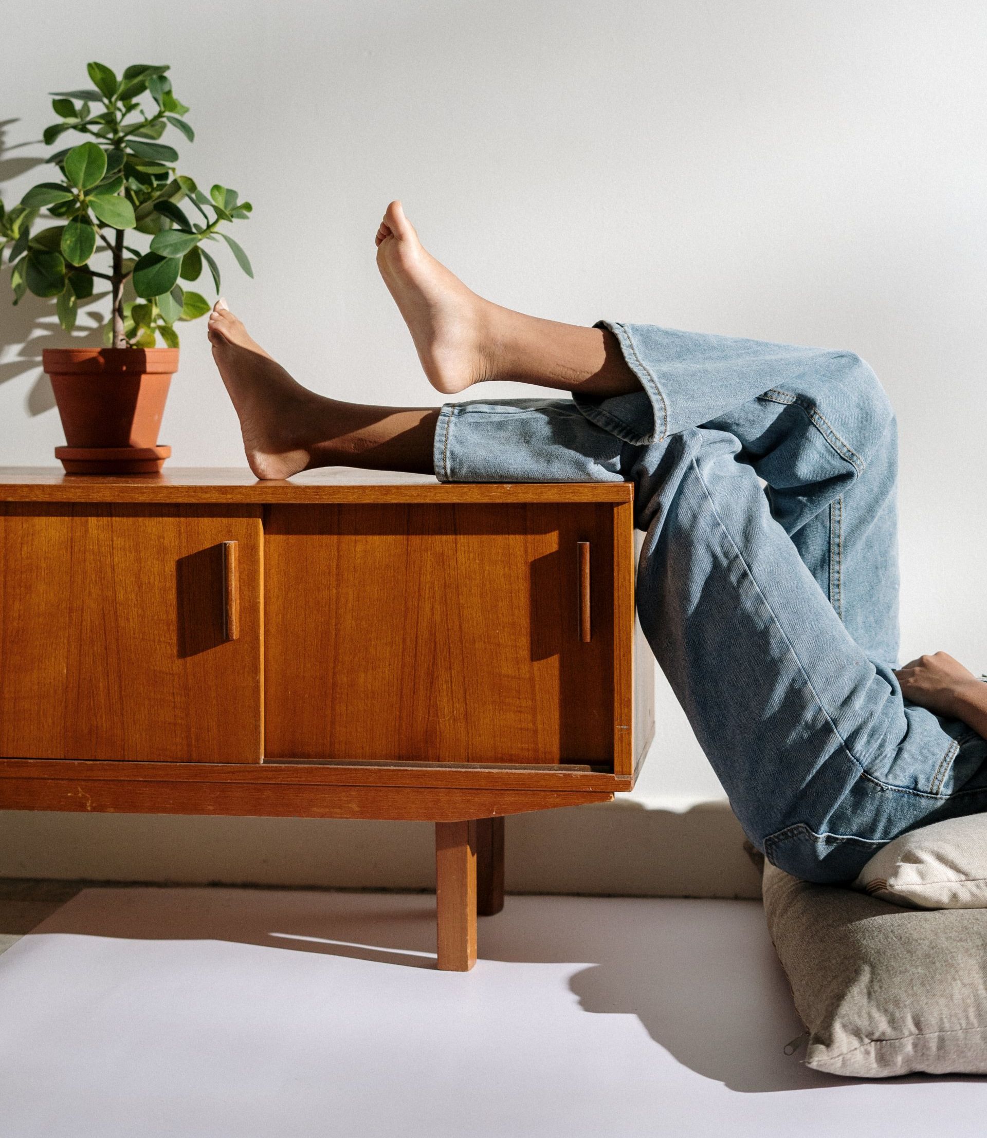 Voeten rustend op een houten kast naast een potplant in een zonnige kamer.