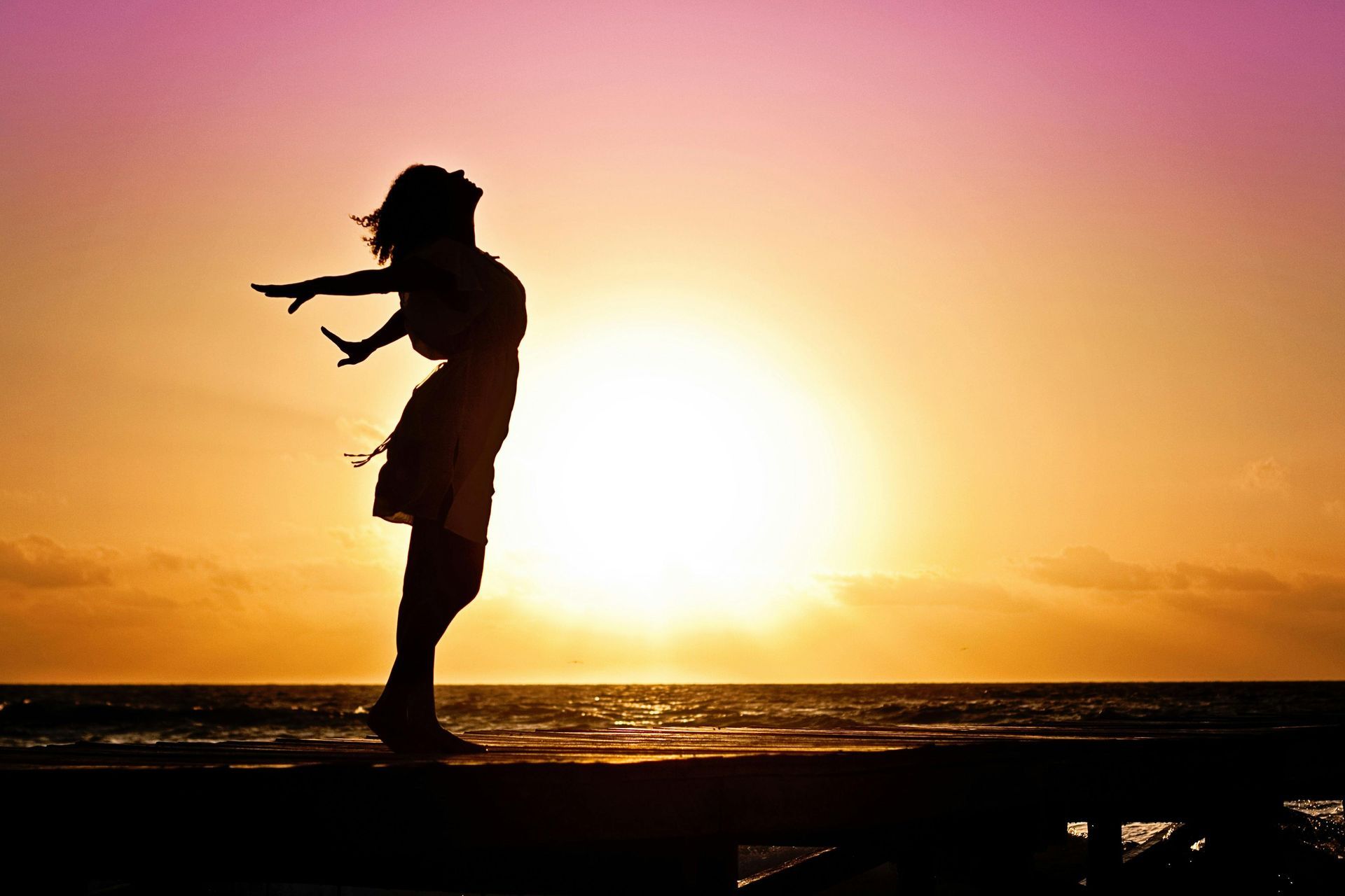 Silhouet van een vrouw met uitgestrekte armen, kijkend naar de ondergaande zon boven de oceaan.