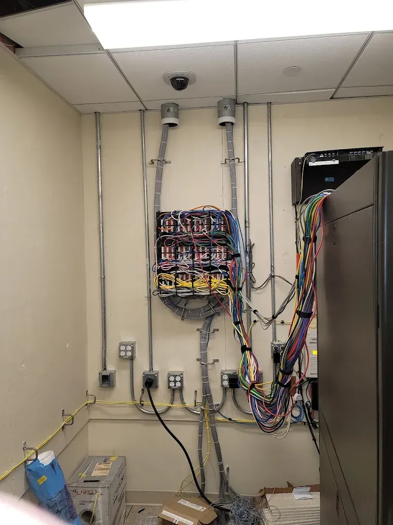 Electrical wiring and equipment mounted on a wall. Conduit, panels, and a server rack.