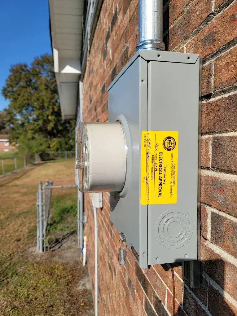 Exterior electrical meter box mounted on a brick wall with conduit; yellow inspection sticker.