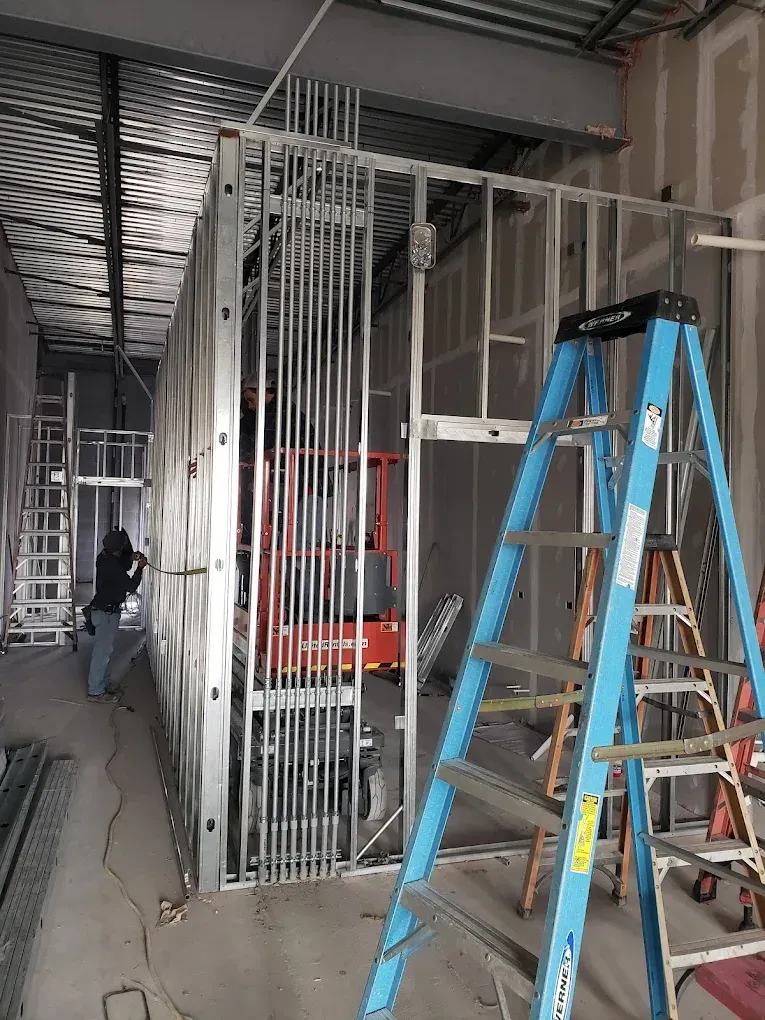 Construction site: Metal studs, conduit, blue ladder, worker.