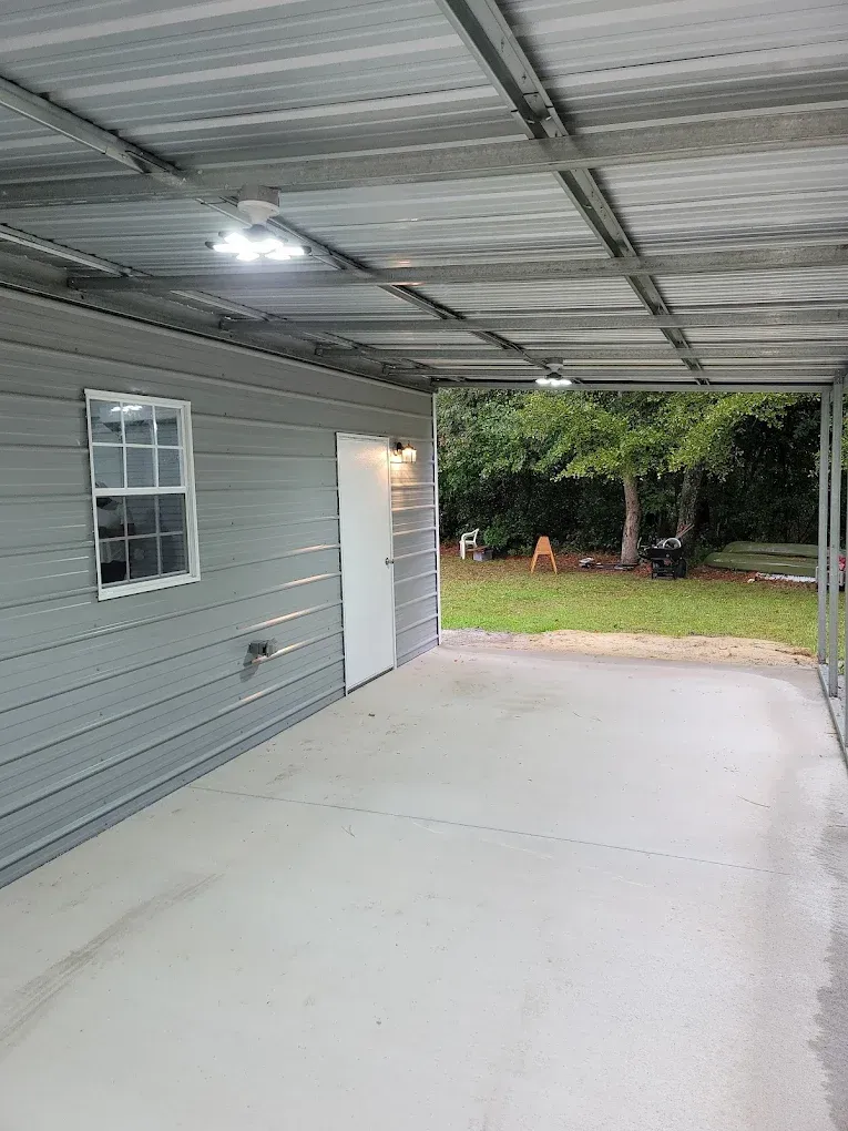 Carport with gray siding, white door, and window. Metal roof, concrete floor, backyard view.