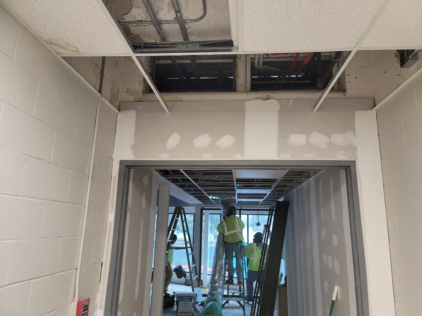 Construction site hallway with workers on ladders, open ceiling tiles, exposed framework, and drywall.