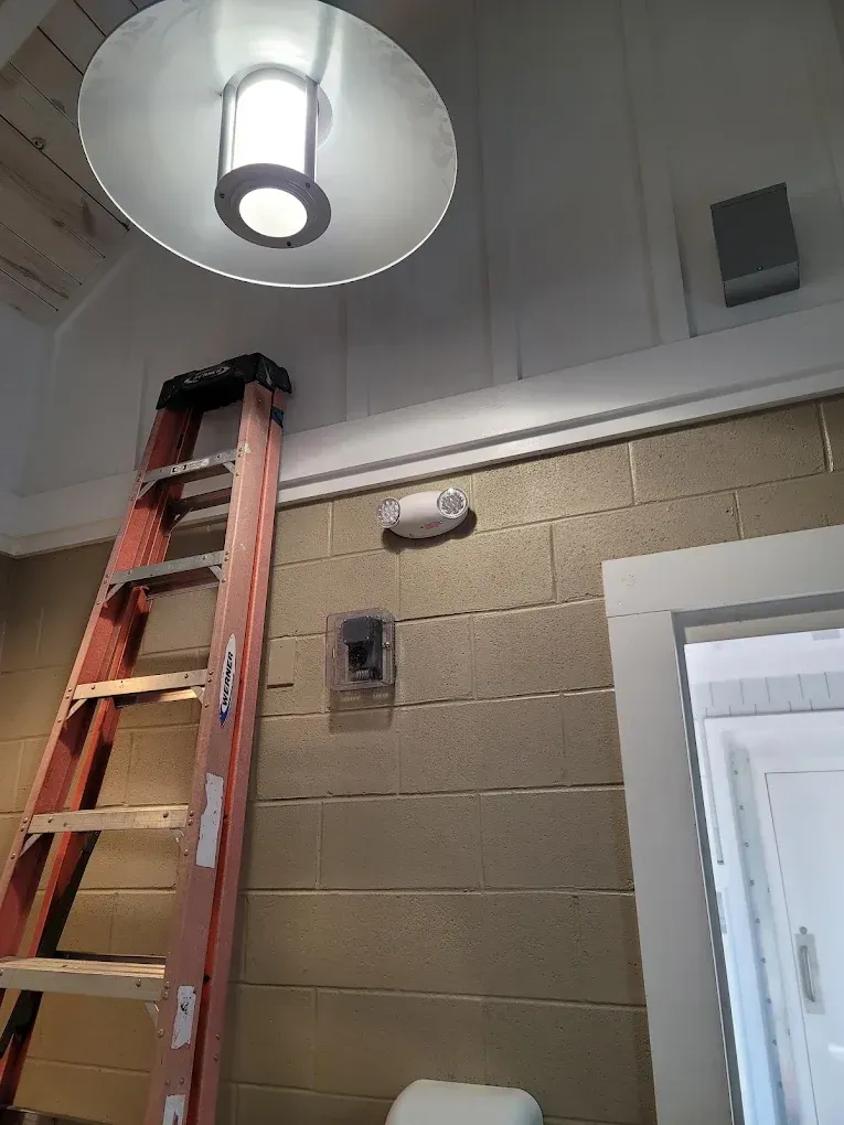 A step ladder next to a tan brick wall with a security camera, a light fixture and a door frame.