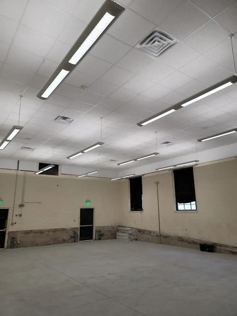 Empty room with linier fluorescent lights on ceiling, windows with black shades, and doors.