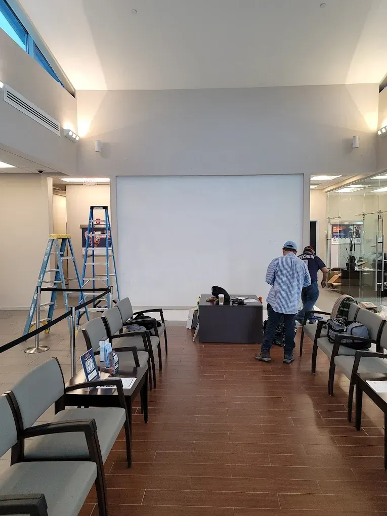 Awaiting area with a large blank screen, chairs, and two people standing near a table with equipment.