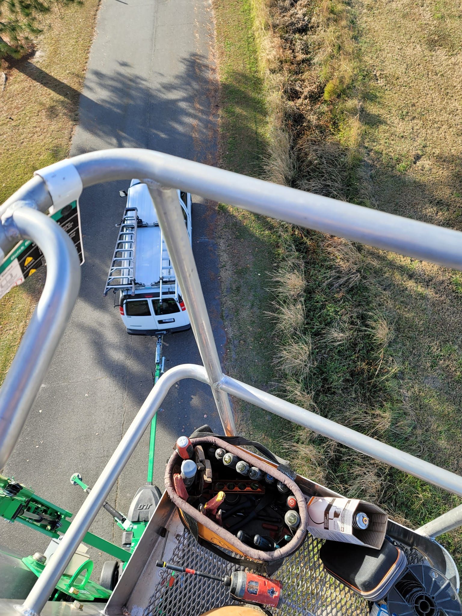 View from lift basket down on service van parked on road. Tool belt, grass, and road in view.