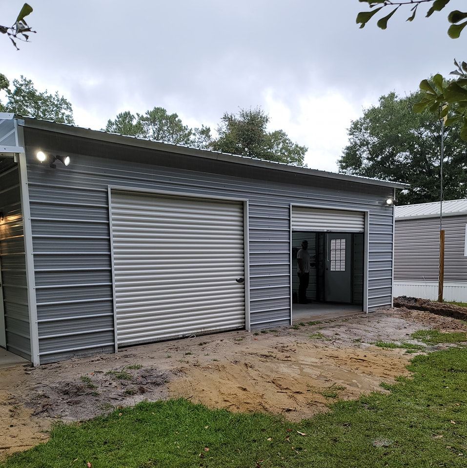 Gray metal garage with two bays, one door open, set on a sandy lot.