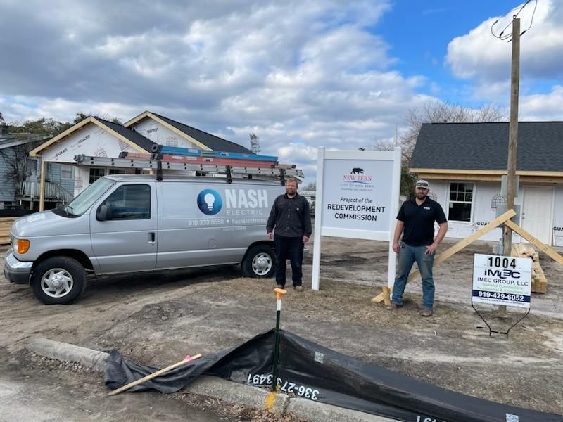 Two men standing by a sign for a construction company. A van and buildings under construction are nearby.