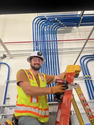 Construction worker smiling, using power tool on ladder, electrical conduit in background.