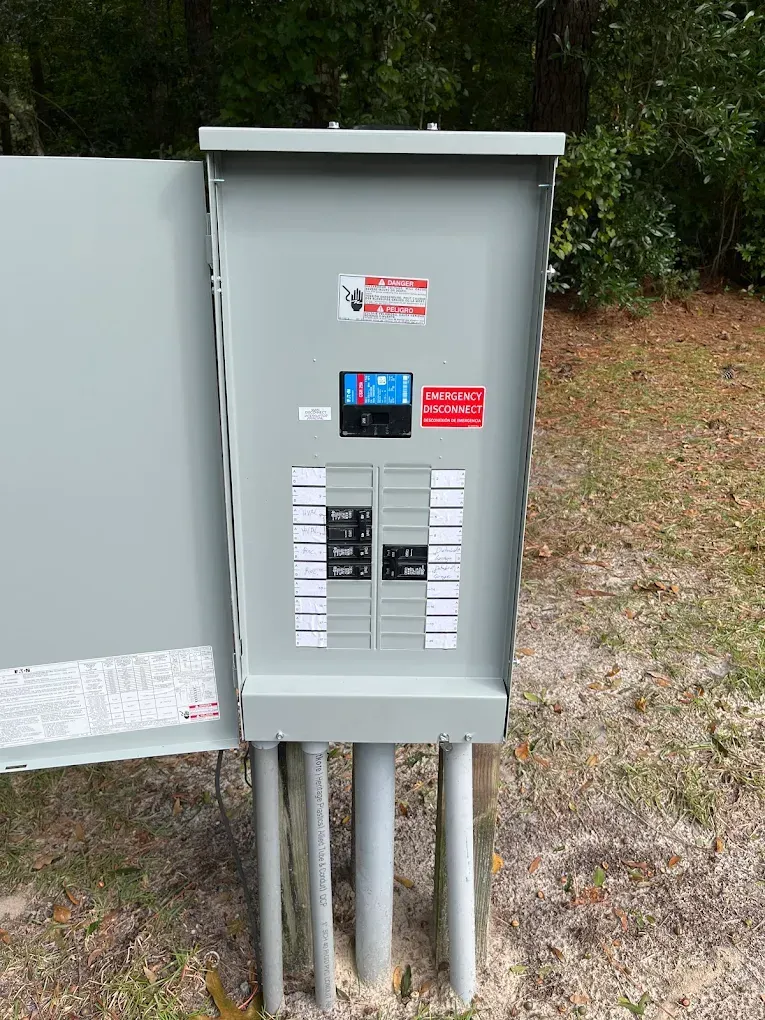 Gray electrical panel outdoors, mounted on posts, with circuit breakers and warning labels.