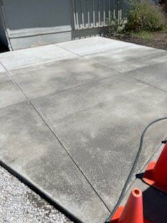 Concrete patio with uneven coloring, bordered by a gravel driveway, safety cones.