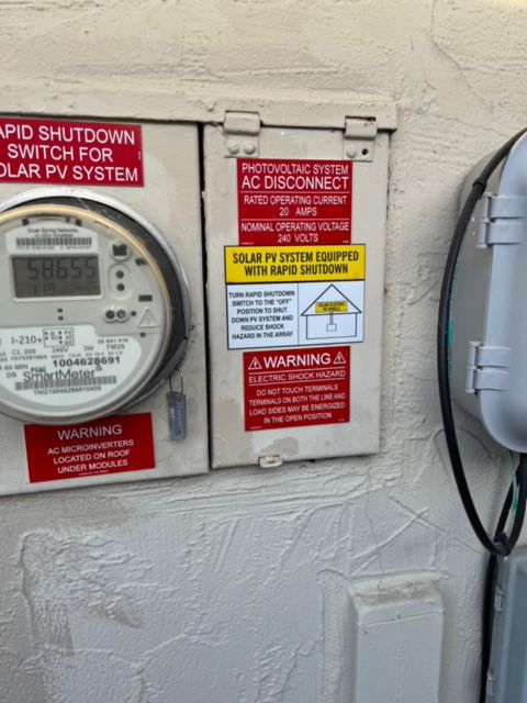 Electric meter, solar panel AC disconnect box with warning labels attached to a stucco wall outside.